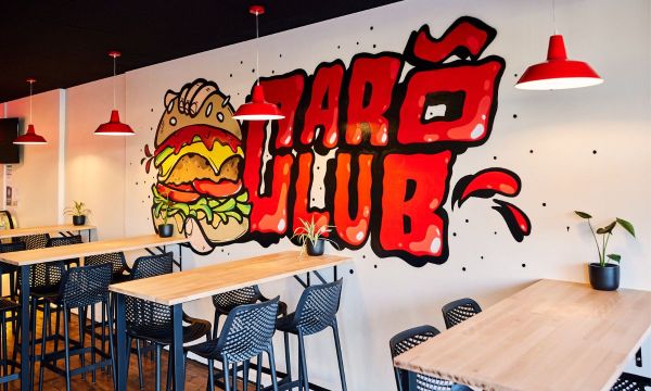 An image of CaroClub Edwardstown's resturant interior, featuring tall tables and black stools lined against a wall that has a graffiti-art style mural with the CaroClub logo showing a hand grasping a large burger. 